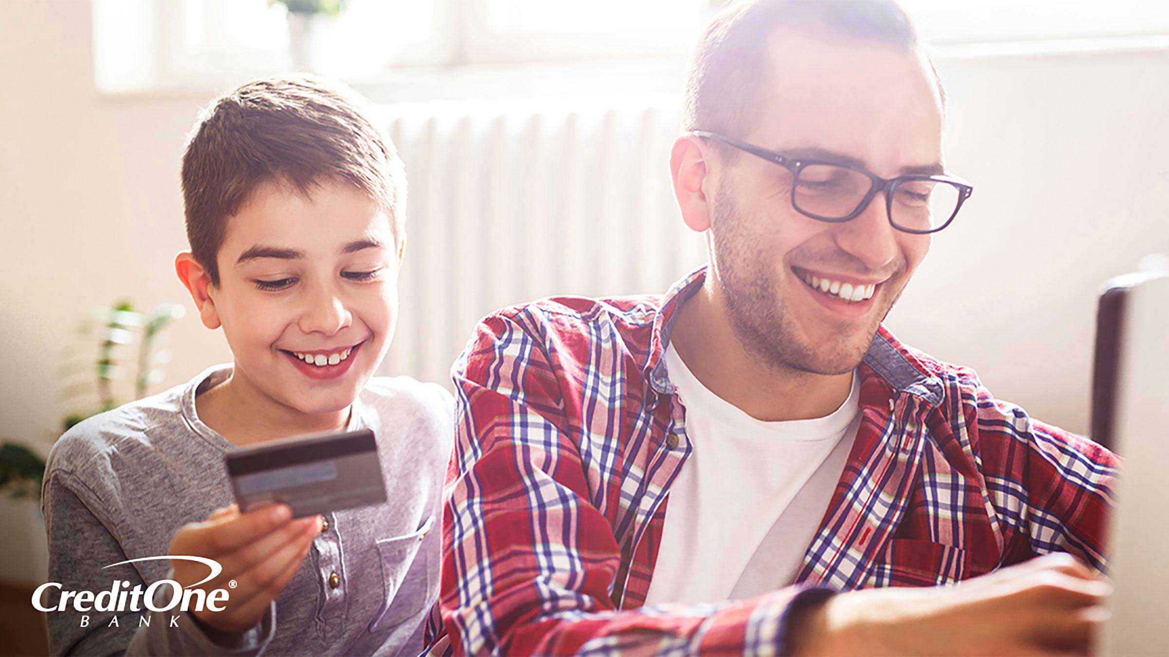 A man types on his computer as his son reads information from a credit card. How to responsibly use a card online is one of many essential credit card lessons for kids.