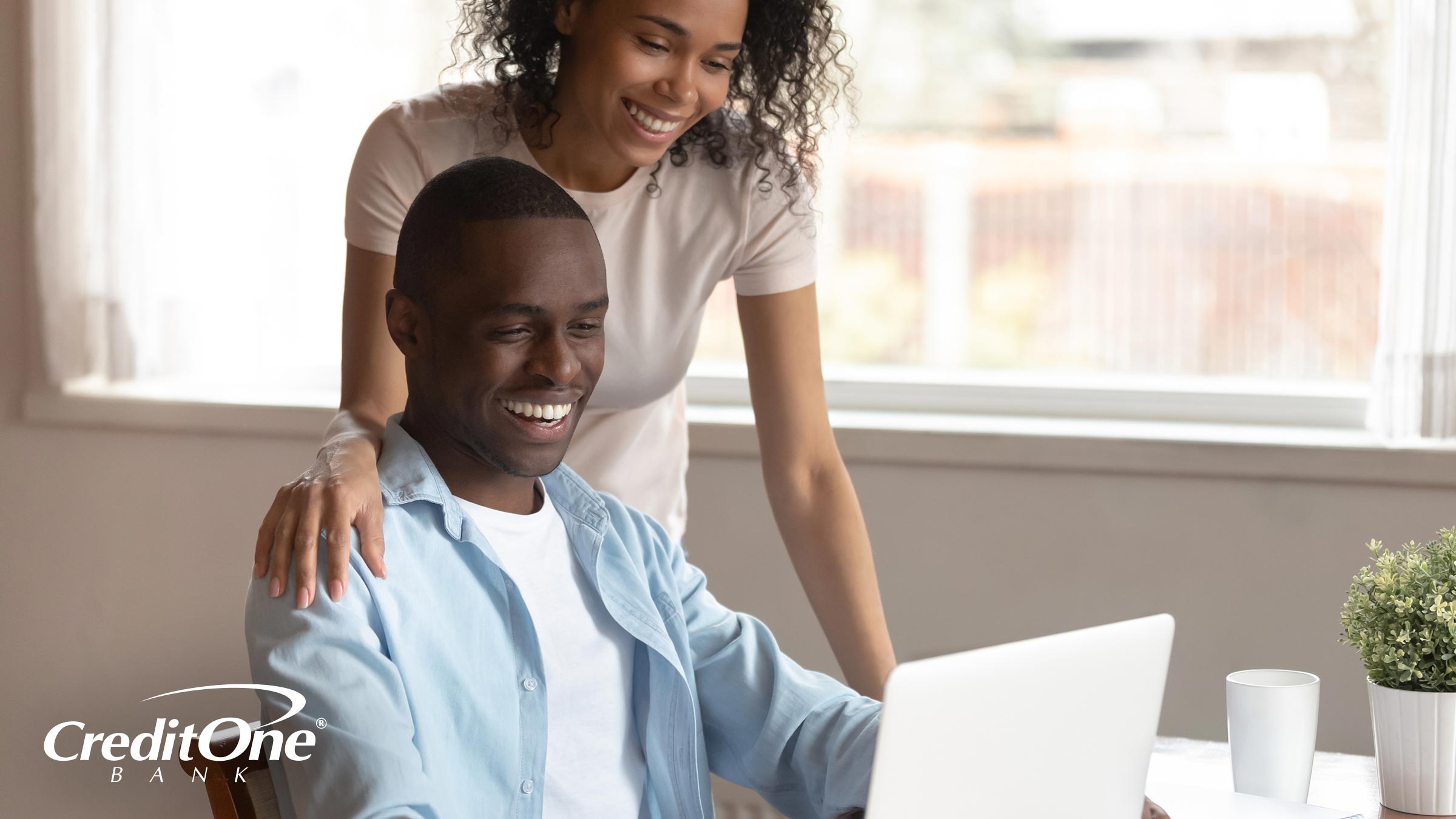 A happy couple smiles as the wife watches her husband working on his laptop. This is how you may react when you redeem rewards from your credit card.