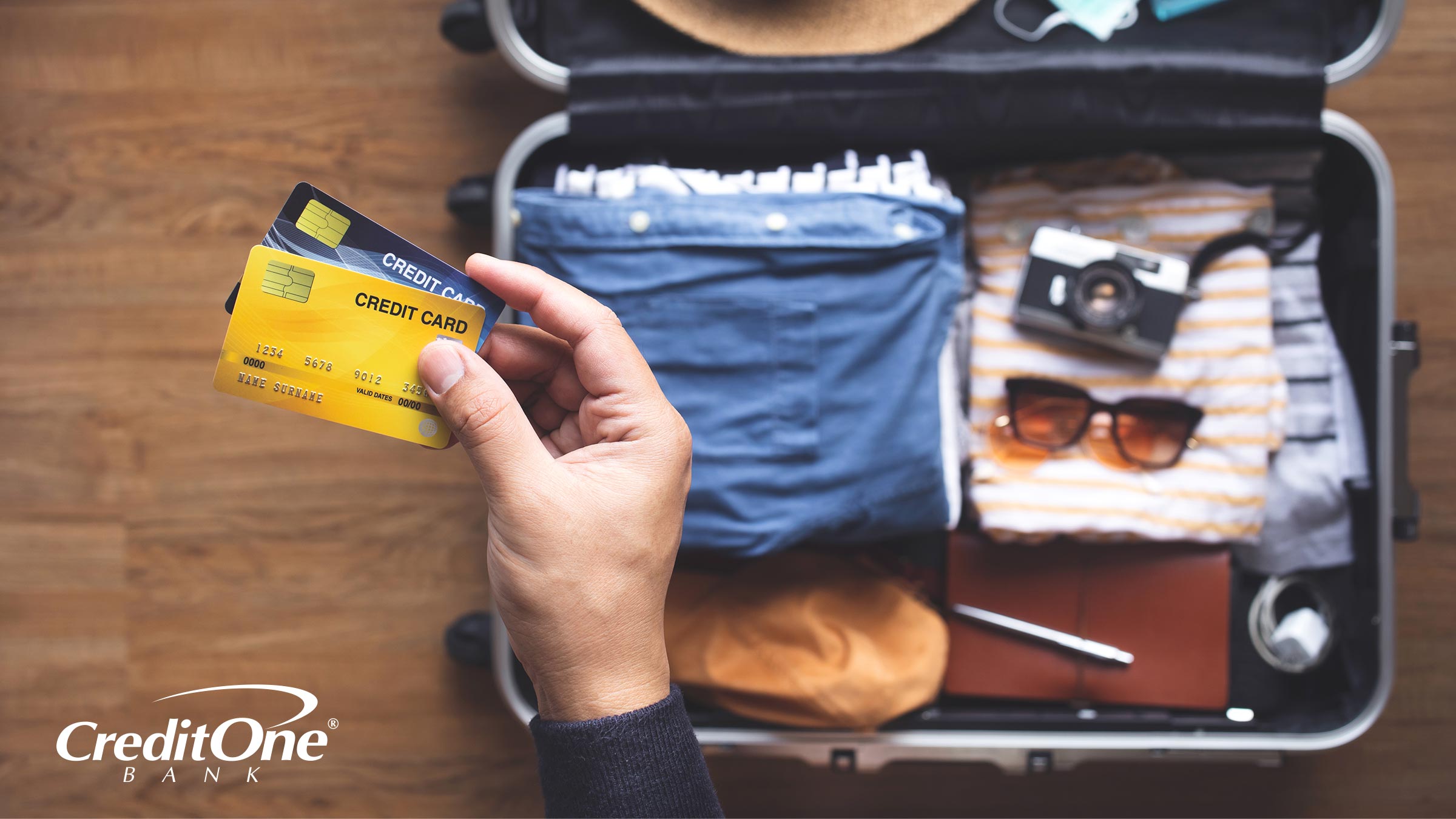 A man’s hand holds two travel credit cards above his suitcase, which is packed and ready for a trip.