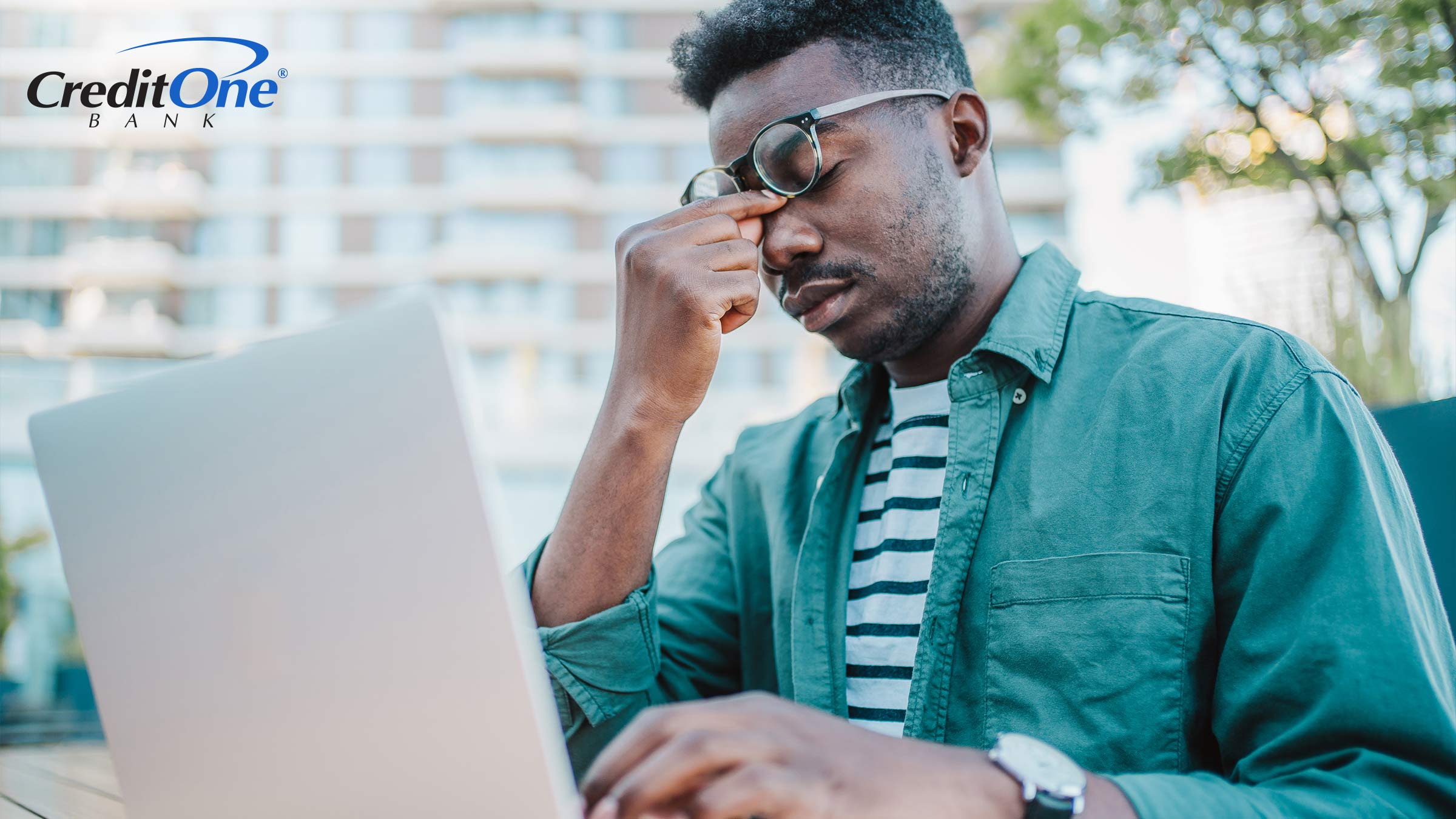 A man sitting at an outside table with his laptop holds the bridge of his nose as he pushes his glasses up and closes his eyes. He is clearly stressed out, which could be from navigating financial challenges and bankruptcy. 