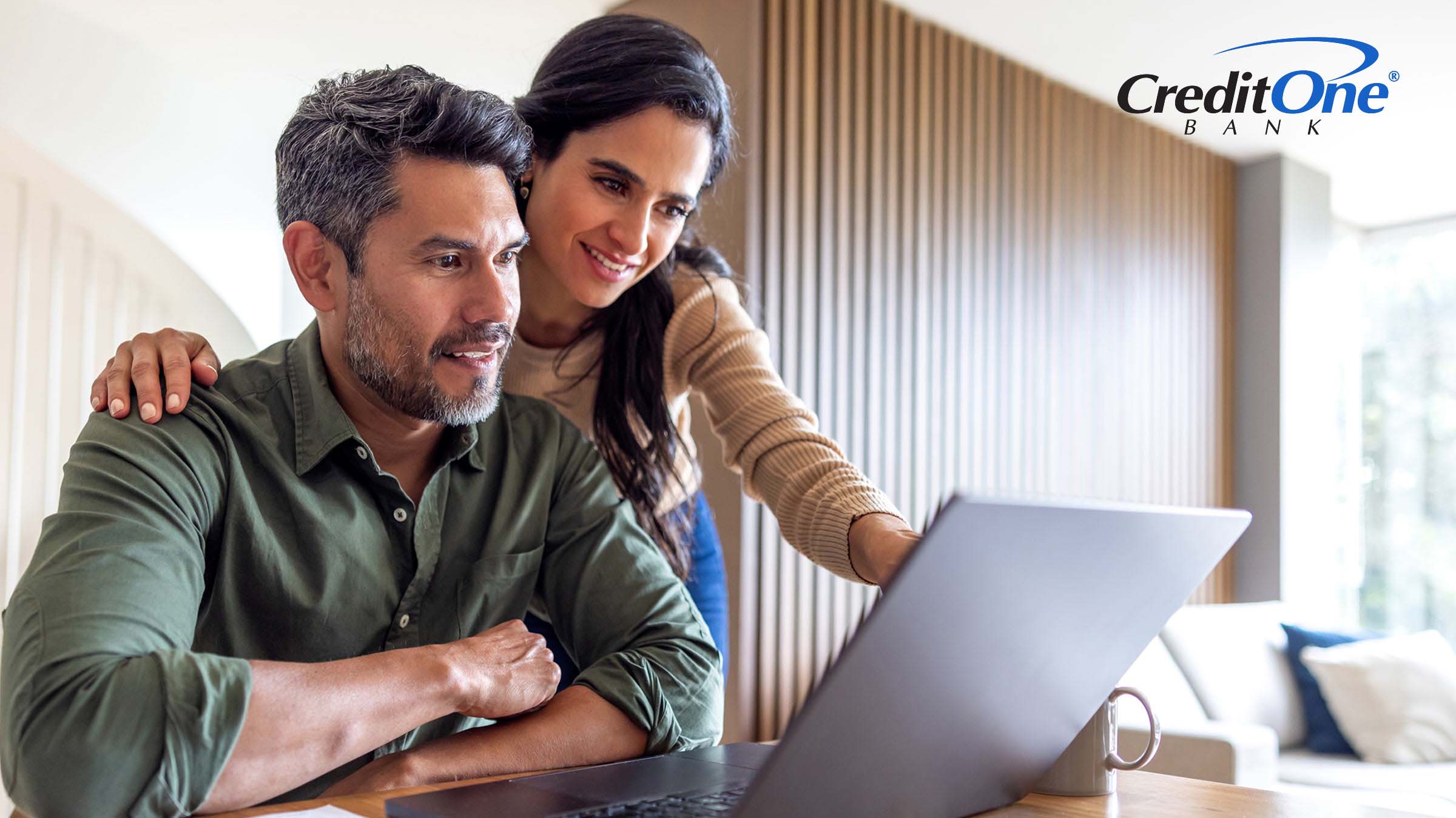 A wife looks over her husband’s shoulder to point at his laptop screen, which is next to some financial statements. They are both smiling, perhaps having found a way to get their budget back on track.