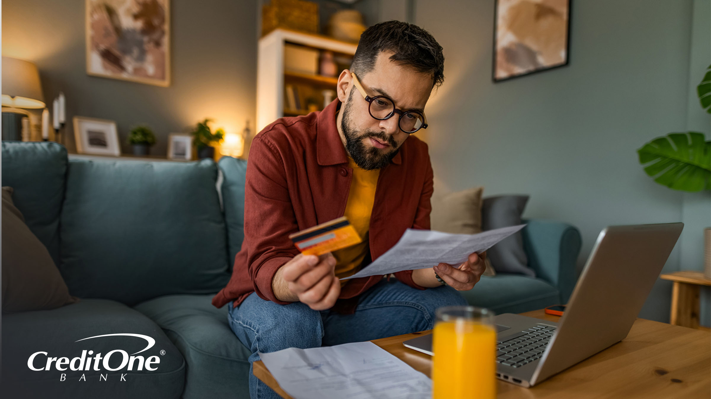 A man sits in his living room, holding up a credit card while reviewing his statement with a concerned expression. He may have noticed something unfamiliar, like a negative balance.